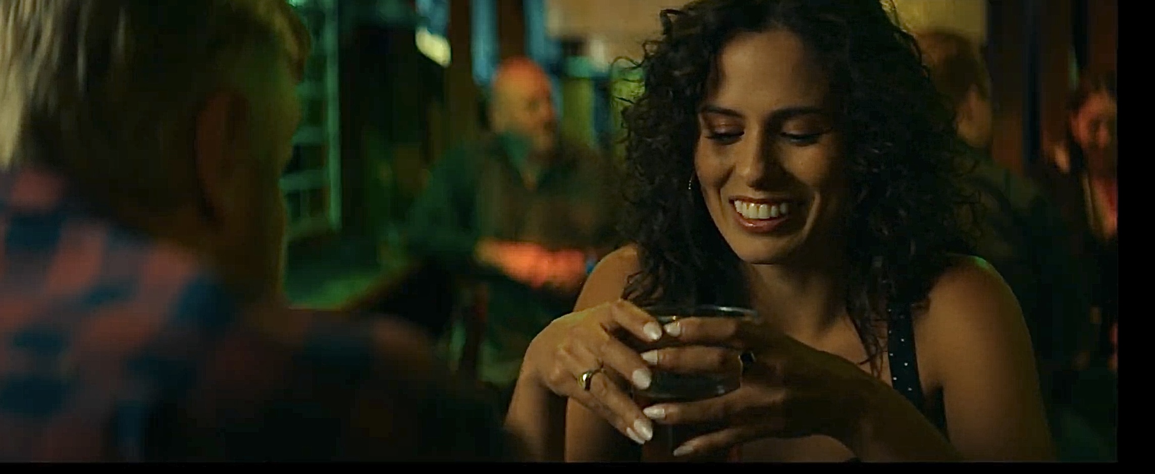 A woman holding a drink in a warmly lit bar, in a still from Other You